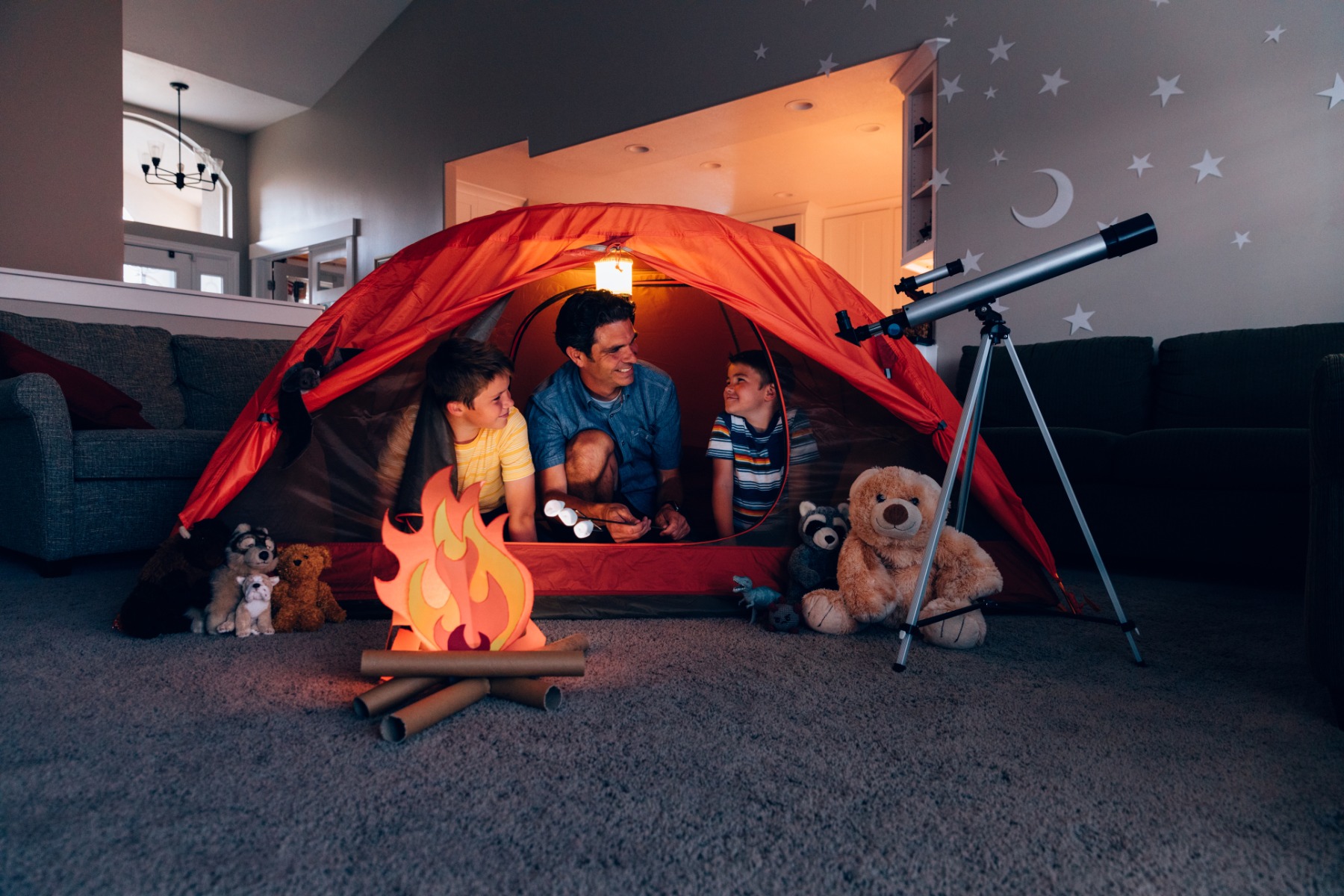 Un père joue avec ses deux fils dans le salon, sous une tente rouge et un feu de camp en carton.